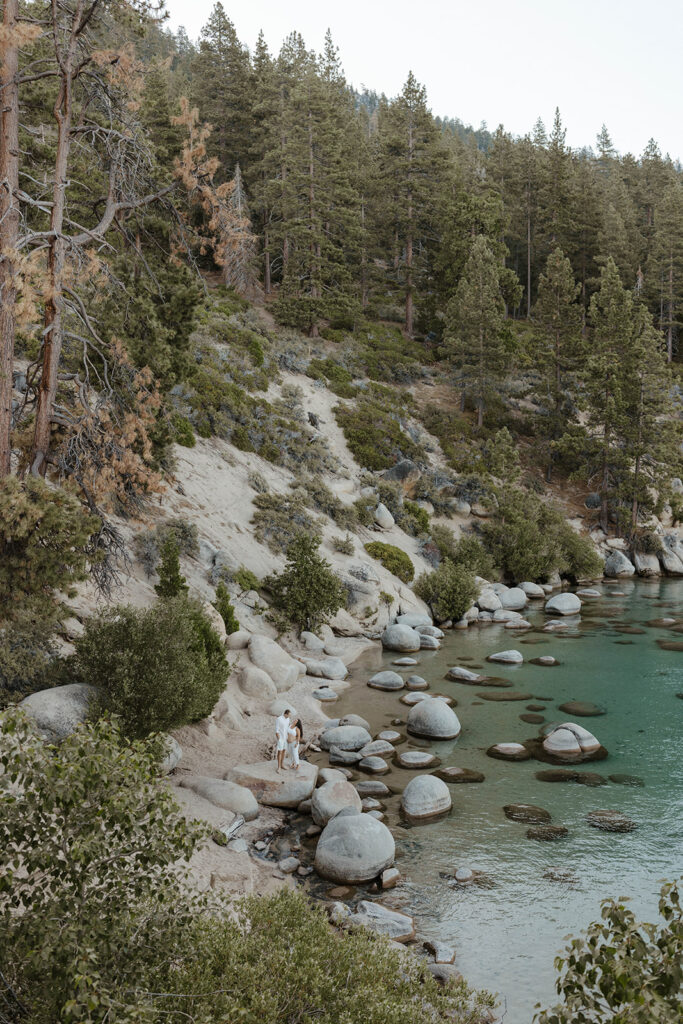 Married couple in distance walking along rocks together on Lake Tahoe beach surrounded by pine trees