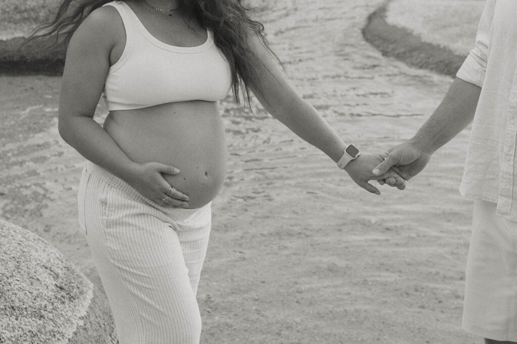 Close up of married couple holding hands on sandy beach in Lake Tahoe while wife holds pregnant belly