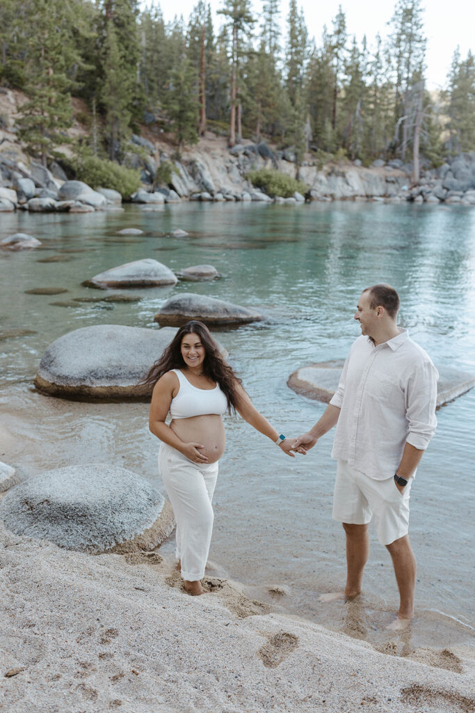 Married couple holding hands while walking barefoot on sandy beach in Lake Tahoe while wife holds pregnant belly and smiles at camera
