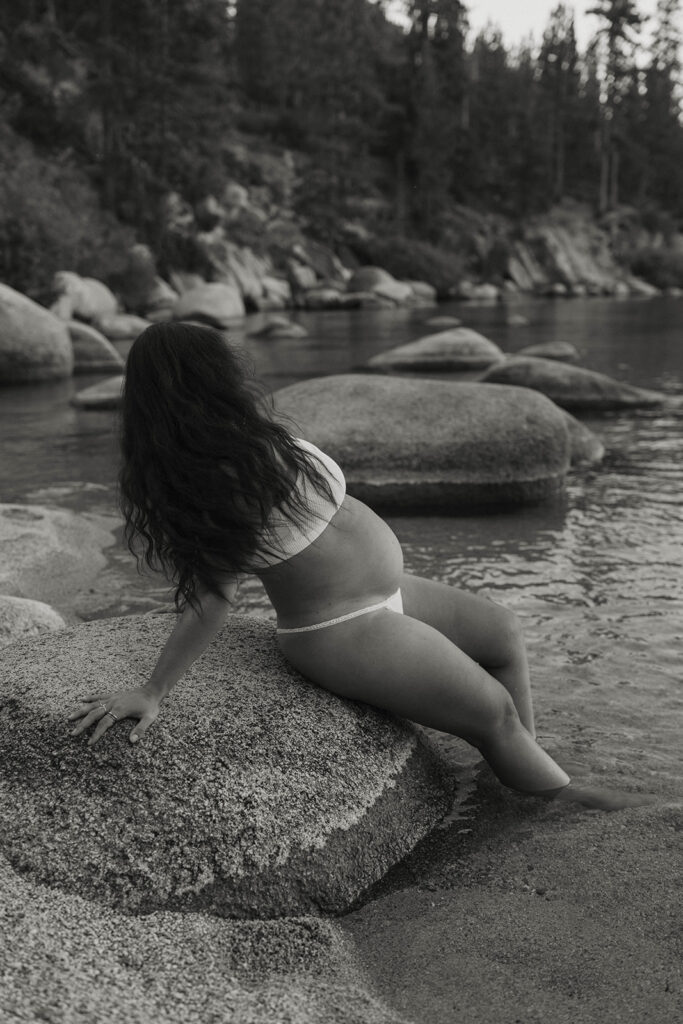 Woman leaning back on rock showing off pregnant belly with feet in the sand on a beach in Lake Tahoe