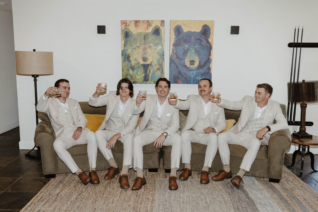 Wedding groom and groomsmen sitting on a couch and holding up drinking glasses together inside at the Landing Tahoe Resort