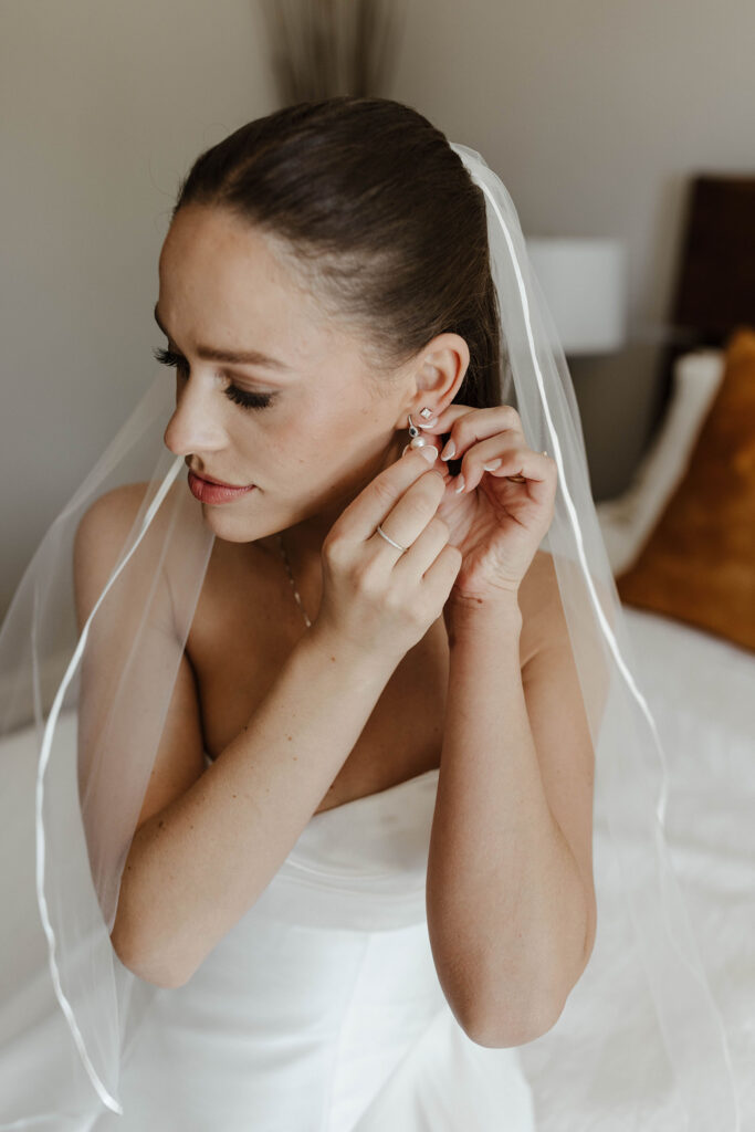 Wedding bride putting on earrings while sitting on bed inside at the Landing Tahoe Resort