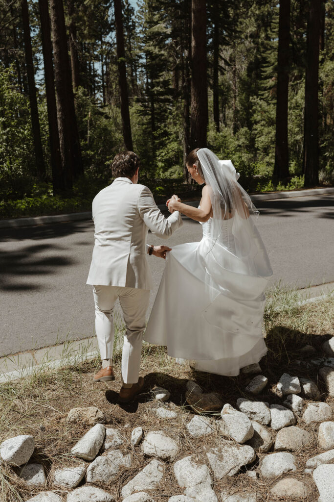 Groom holding wedding bride's hand and dress while walking over rocks towards street and pine trees at the Landing Tahoe Resort