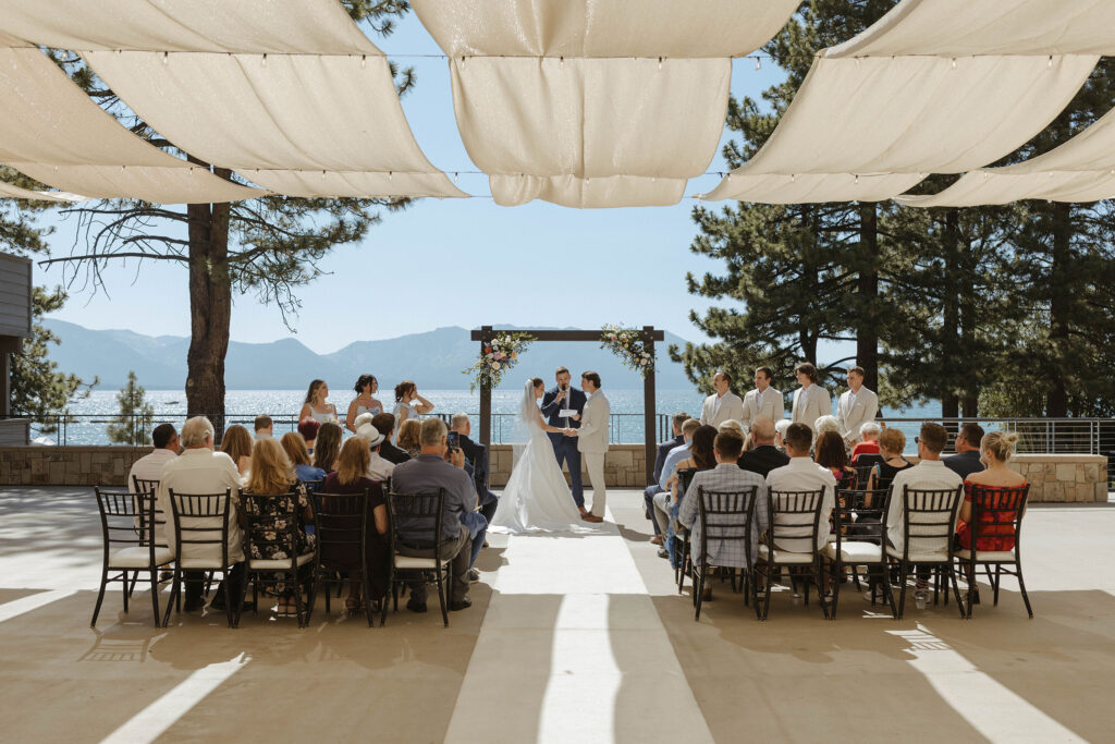 Overview of wedding ceremony with wedding couple holding hands under arch and white cloth draped across string lights above at the Landing Tahoe Resort