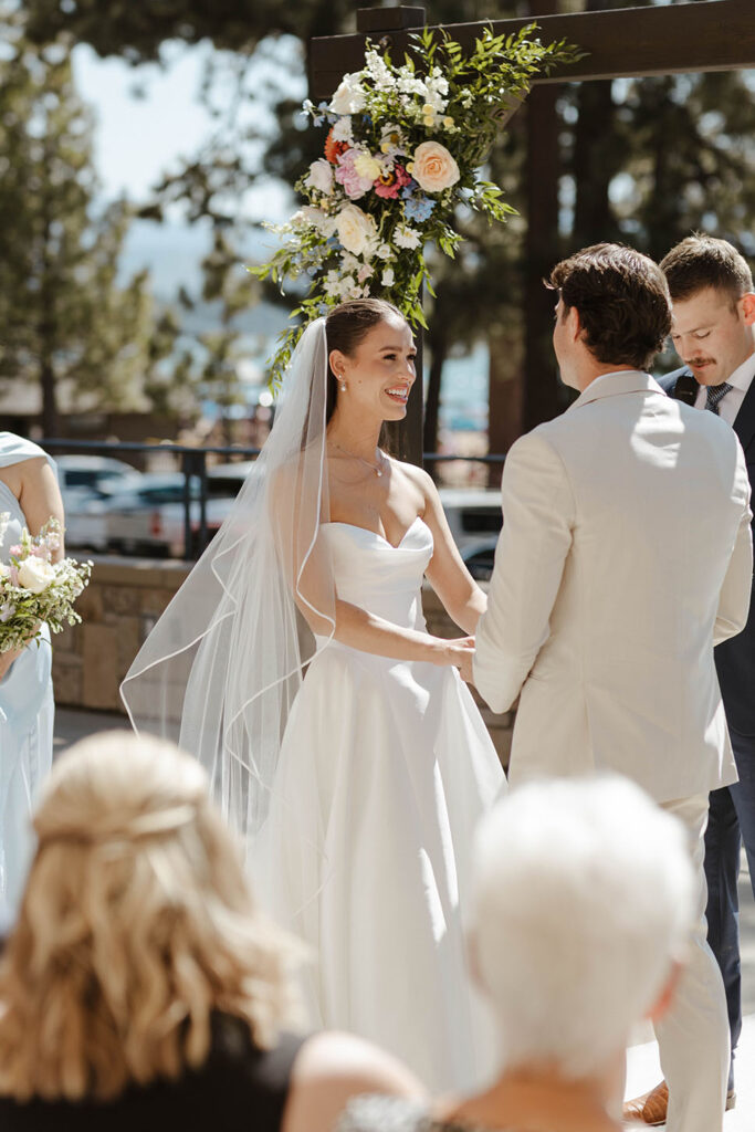 Bride smiling while holding grooms hand during wedding ceremony with colorful flowers decorating arch behind her at the Landing Tahoe Resort