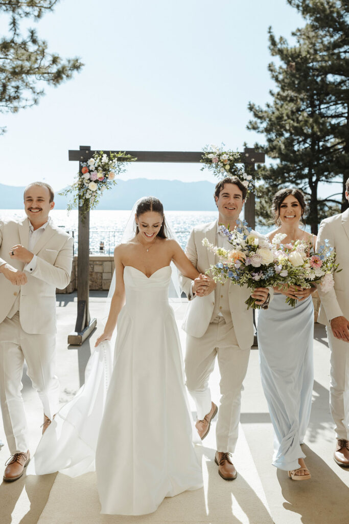 Wedding couple holding hands and smiling while walking out of ceremony with bridal party at the Landing Tahoe Resort with arch and Lake Tahoe in background