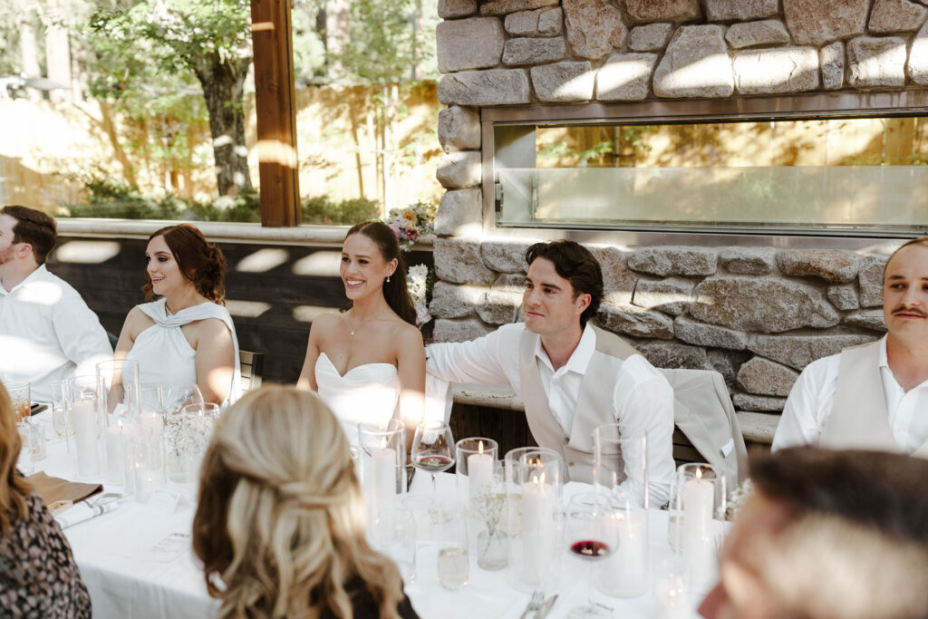 Groom with his arm around bride while they are sitting at wedding reception table and listening to speeches with rock pillar behind them at the Landing Tahoe Resort