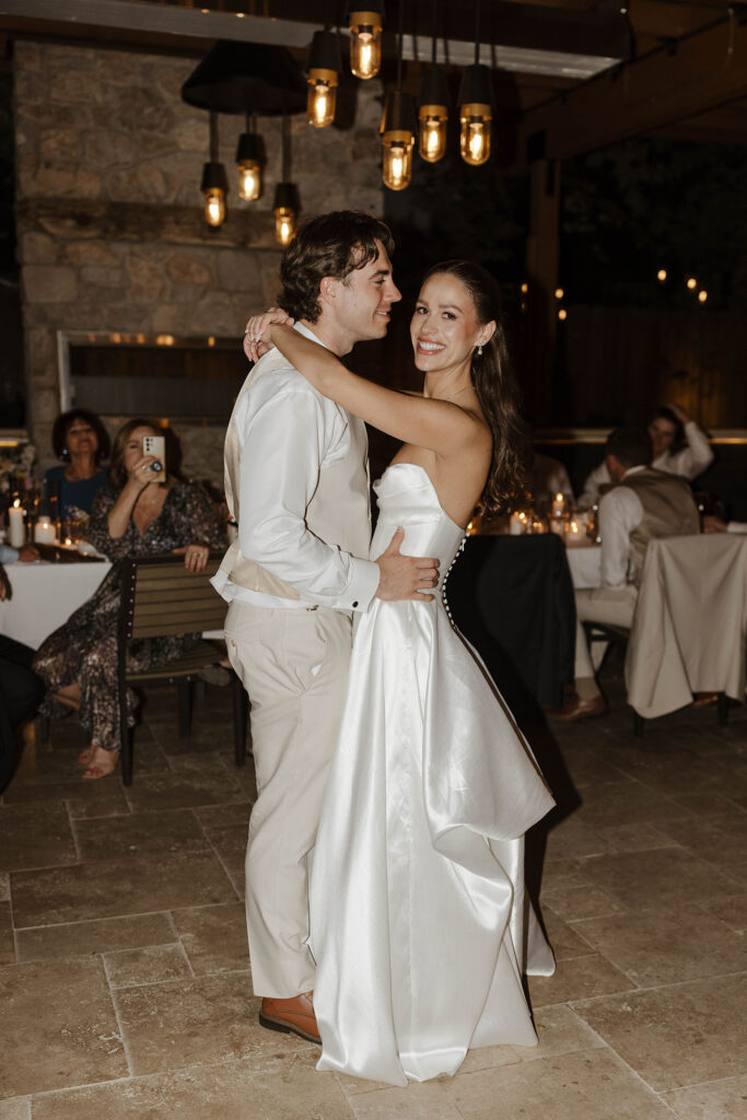 Bride smiling at camera during couple's first dance while groom holds her and warm lights hang above them at the Landing Tahoe Resort