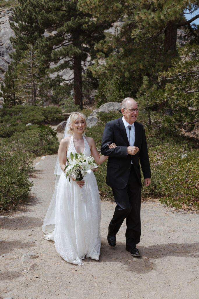 Bride holding dad's arm while walking down to outdoor elopement ceremony in Lake Tahoe with pine trees and scenery in background