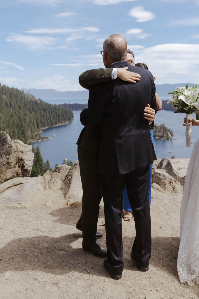 Groom hugging bride's dad during elopement ceremony with Lake Tahoe and pine trees in background