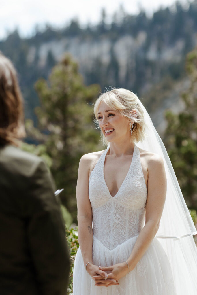 Bride smiling while listening to groom read vows during elopement ceremony in Lake Tahoe with pine trees in background