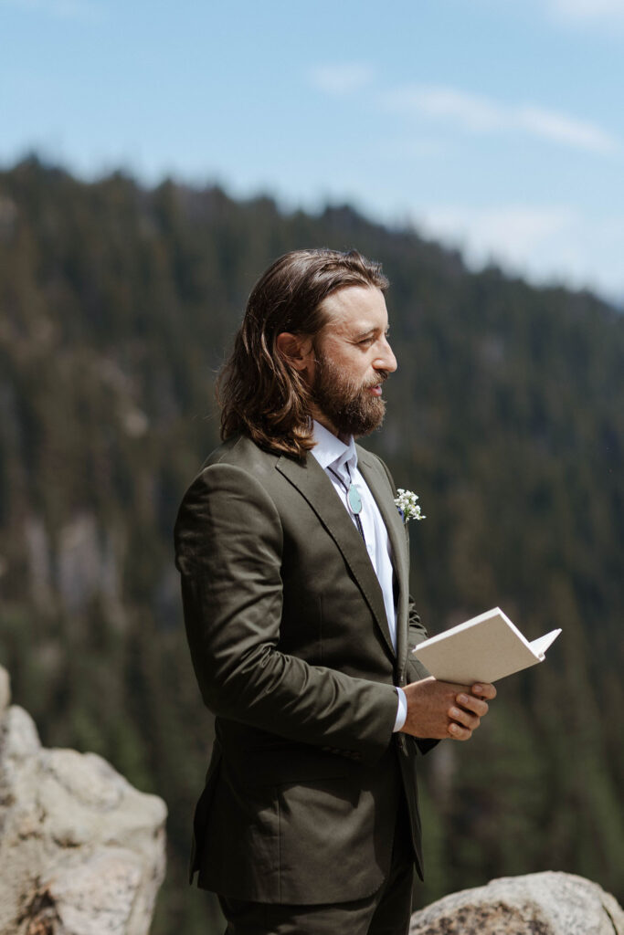 Groom reading vows from vow book during elopement ceremony in Lake Tahoe with pine trees in background