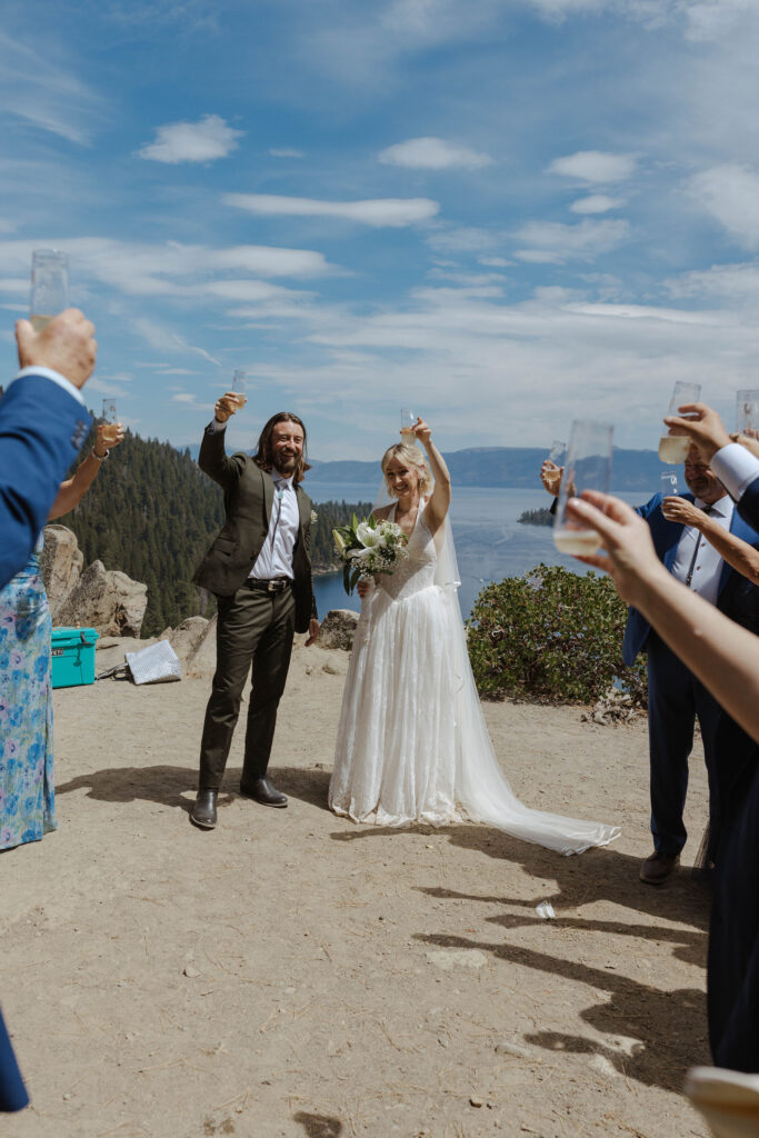 Elopement couple smiling and holding champagne glasses up with guests in celebration after ceremony in Emerald Bay with Lake Tahoe in background
