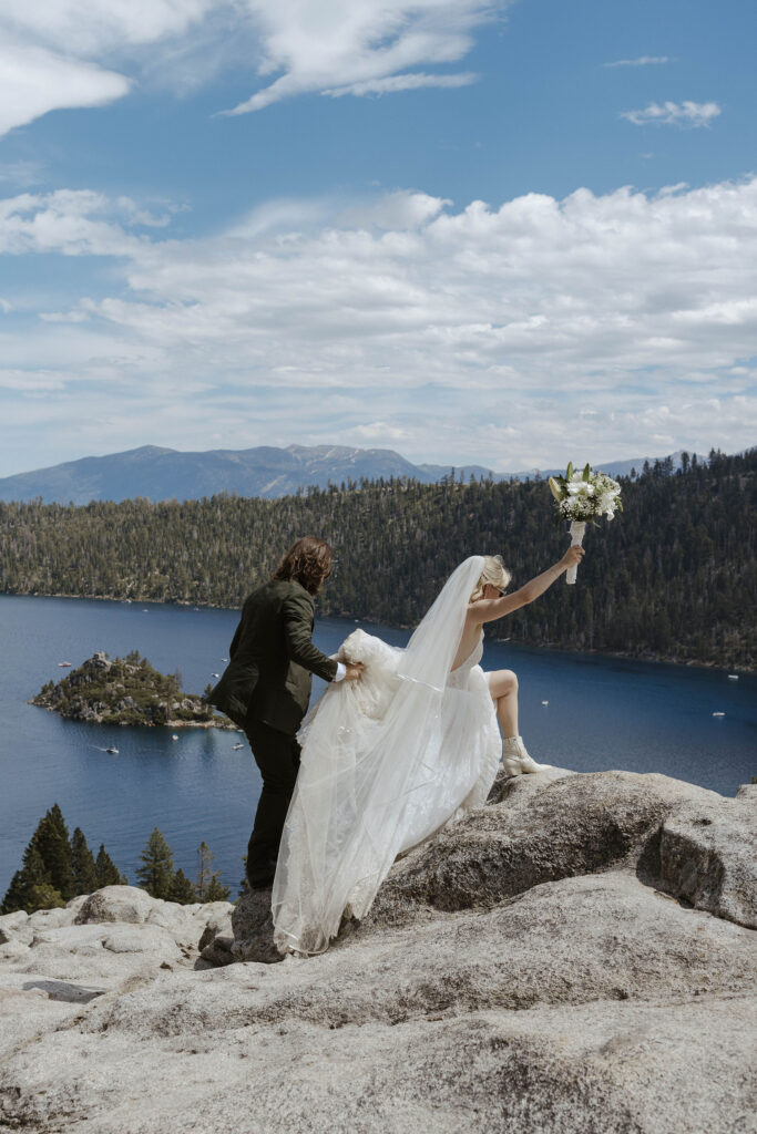 Elopement bride stepping up on rock while holding floral bouquet in the air and groom holds bride's dress in Lake Tahoe with Emerald Bay in background