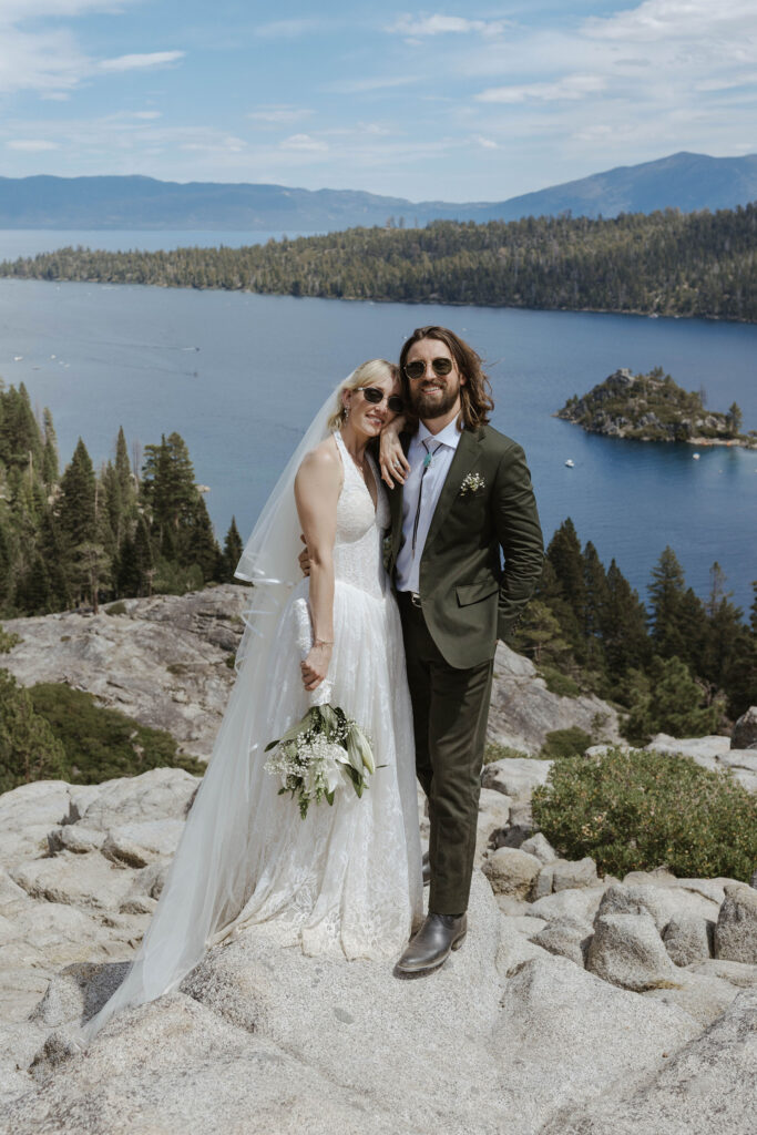 Elopement couple standing together and smiling at camera with sunglasses on while bride leans on groom's shoulder in Lake Tahoe with Emerald Bay in background
