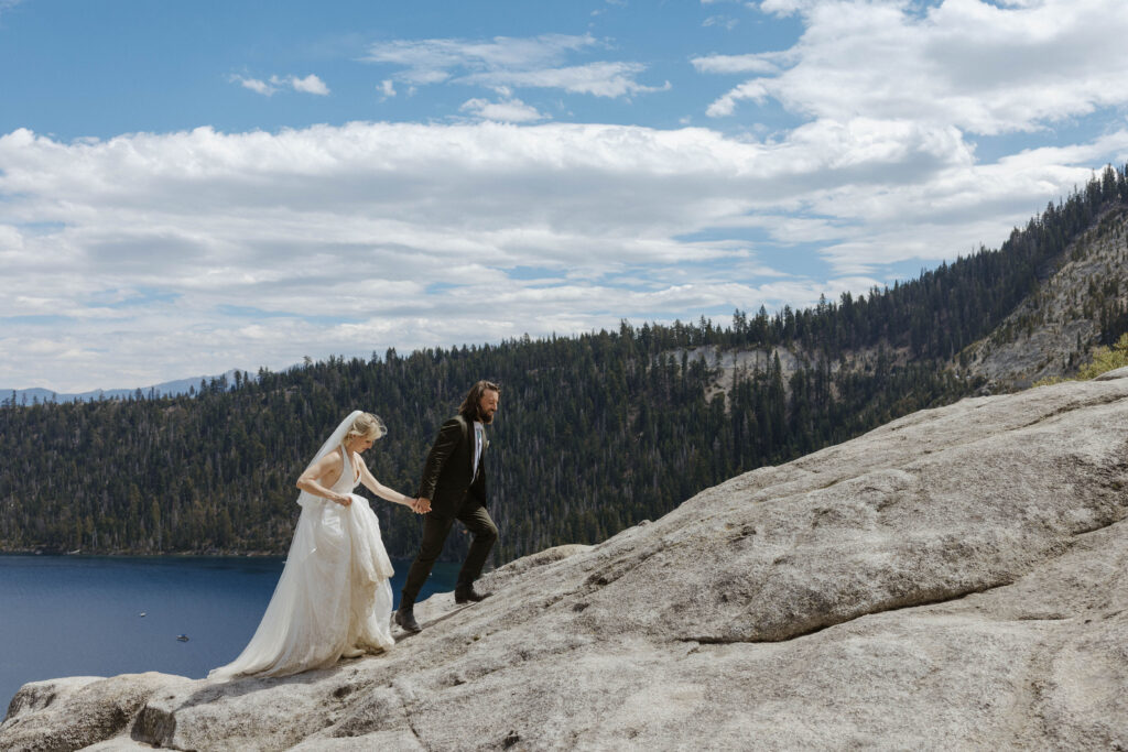 Elopement groom holding bride's hand while leading her up large rocky hill in Lake Tahoe with lots of pine trees in background and cloudy skies above them