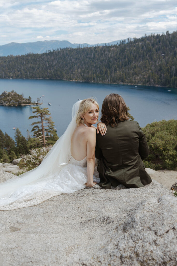Elopement couple sitting on large rock hill together while bride looks back over shoulder at camera and smiles in Lake Tahoe with Emerald Bay in background