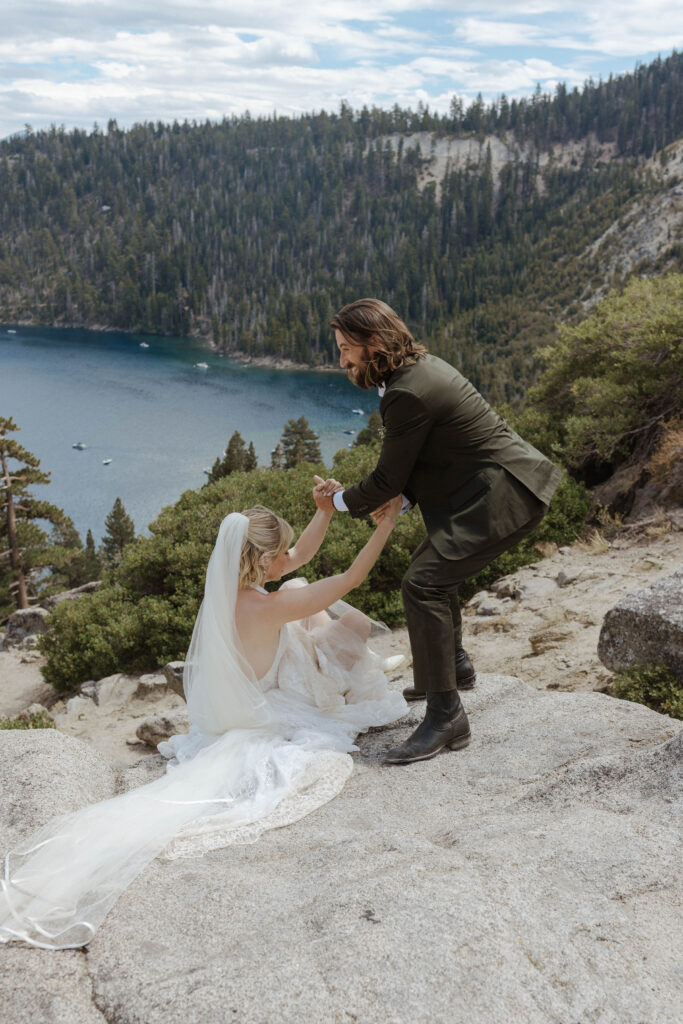 Groom smiling while helping elopement bride get up from rocky hill in Lake Tahoe with pine trees and Emerald Bay in background