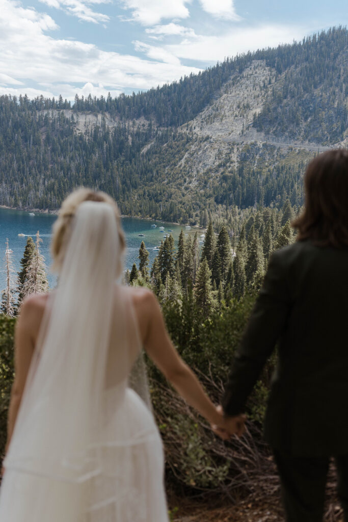 Elopement couple standing and holding hands together while looking out at Emerald Bay and mountainside of pine trees in Lake Tahoe
