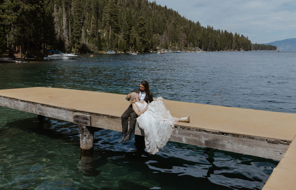 Elopement bride laying on groom's lap while looking at each other and sitting on a pier in Lake Tahoe with pine trees in background