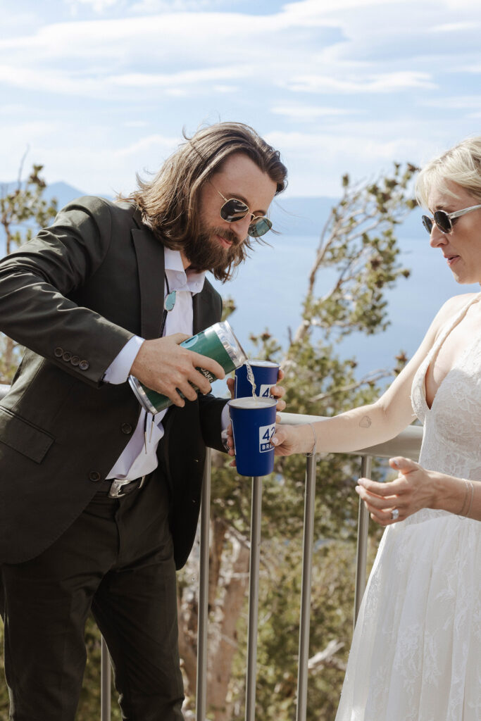 Groom pouring beer into bride's cup while both are wearing sunglasses with Lake Tahoe in background