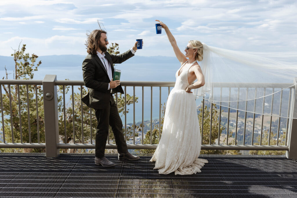 Elopement couple holding cups up together and smiling at each other in front of railing with view of Lake Tahoe in background