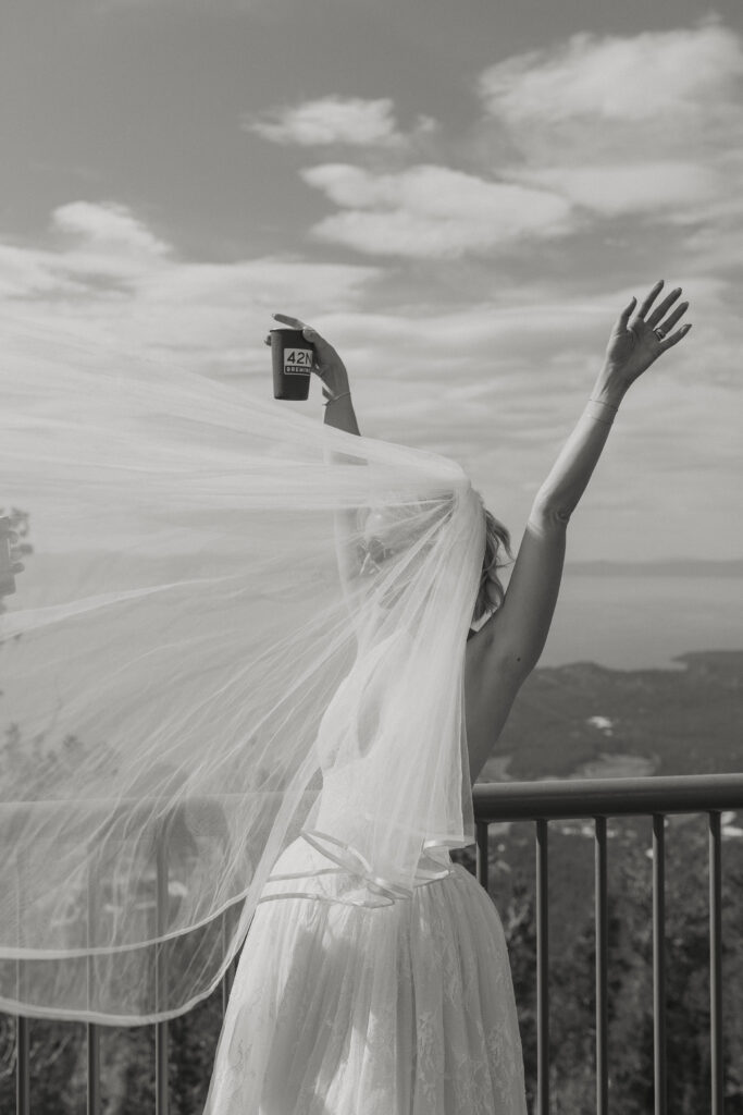 Elopement bride with her arms in the air while holding cup and wind blows veil over her face and Lake Tahoe in background
