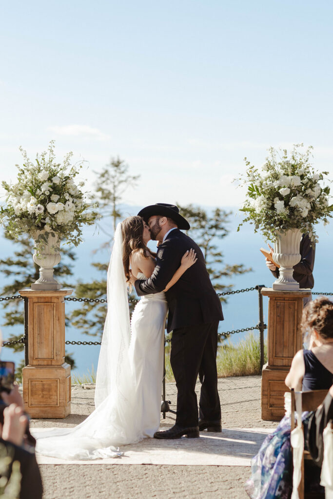 Wedding couple's first kiss during ceremony in between two wooden columns with white flowers at Heavenly Lake View Lodge