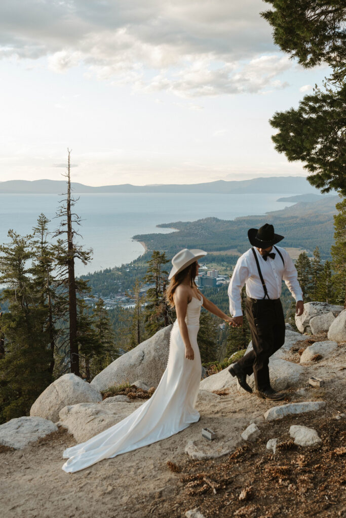 Wedding couple holding hands and walking up trail together while wearing cowboy hats with Lake Tahoe in background at Heavenly Lake View Lodge