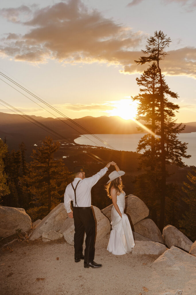 Groom twirling bride on a plateau in front of large rocks while wearing cowboy hats at sunset with pine trees and Lake Tahoe in background at Heavenly Lake View Lodge