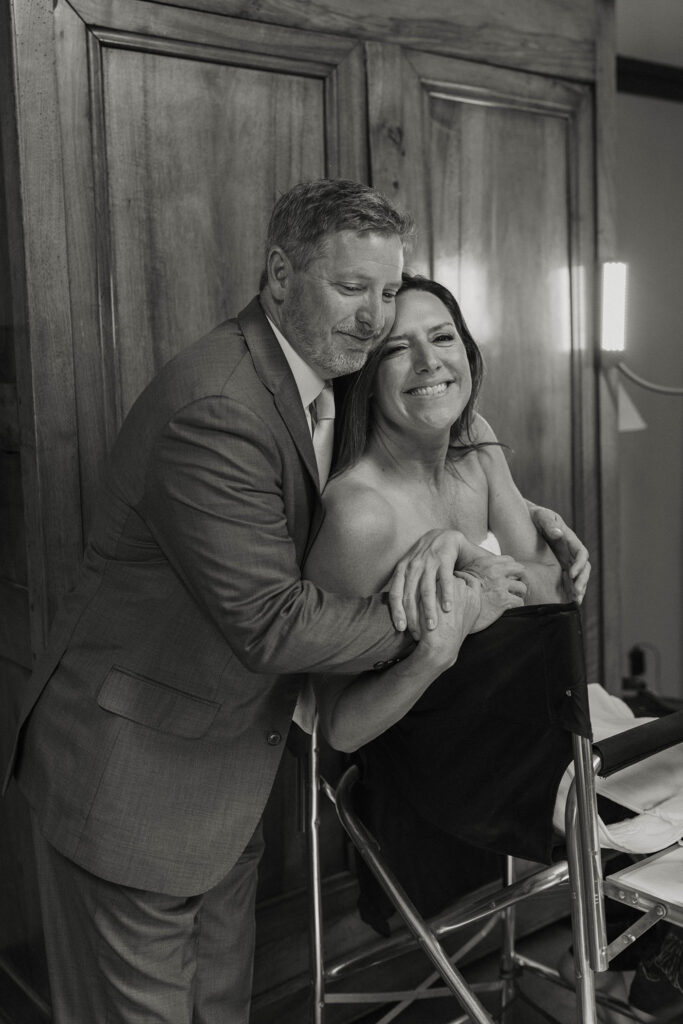 Wedding groom hugging bride sitting in chair while she smiles inside at the PlumpJack Inn