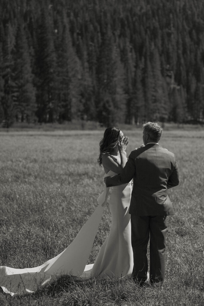 Wedding bride emotional while groom holds arm around her standing in a field together with lots of pine trees in background