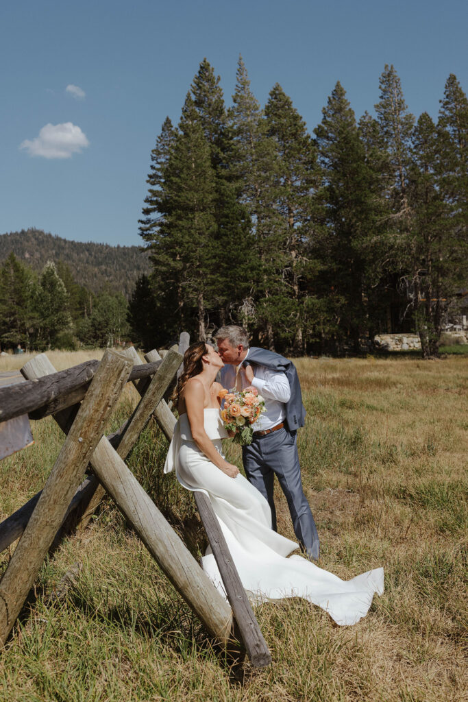 Wedding couple kissing in a field while bride sits on wooden fence and groom holds jacket over shoulder with pine trees in background