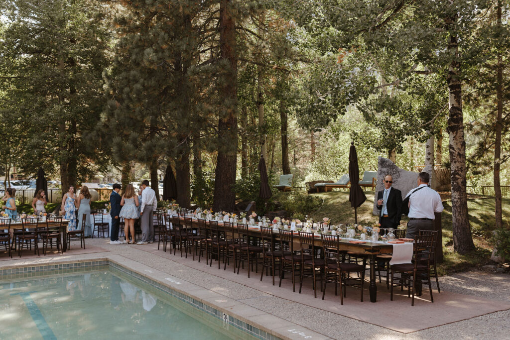 Wedding guests talking with each other while standing around dark wooden reception tables at PlumpJack Inn with tall pine trees in background