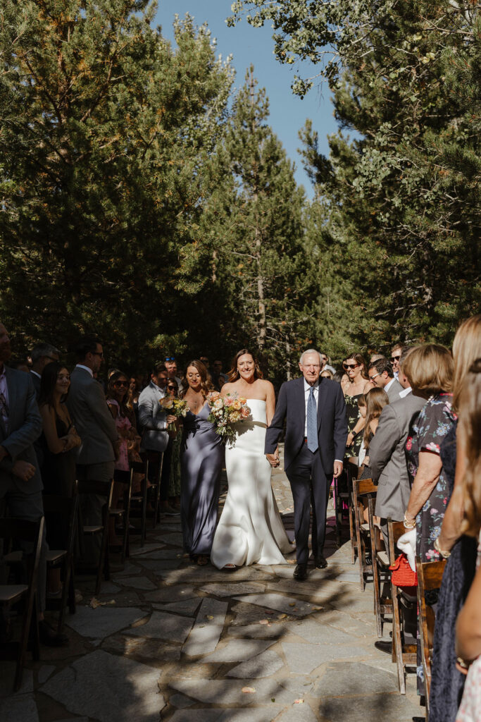 Bride holding dad's hand while walking down wedding aisle to ceremony at the PlumpJack Inn with guests on either side and pine trees in background