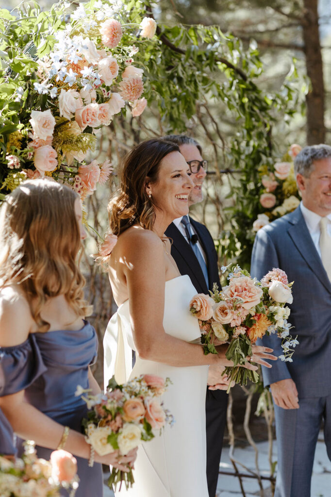 Bride smiling during wedding ceremony while standing in front of arch with lots of greenery and colorful flowers and holding colorful floral bouquet at the PlumpJack Inn