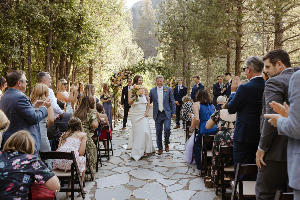 Wedding couple walking down aisle together after ceremony at the PlumpJack Inn with guests clapping on either side and tall pines and arch with lots of greenery and colorful flowers in background