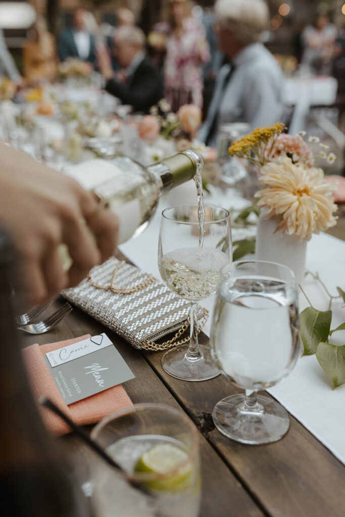 Close up of wine pouring into wine glass on dark wooden reception table with colorful florals and greenery on table at the PlumpJack Inn