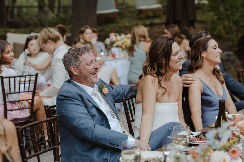 Wedding couple sitting together at reception table and holding hands while smiling and listening to speeches at the PlumpJack Inn