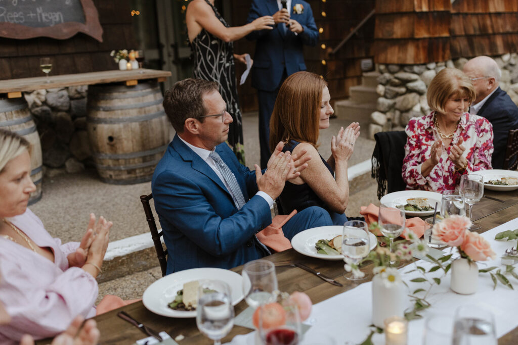 Wedding guests clapping while sitting at reception table listening to speeches with white tableware and colorful florals at the PlumpJack Inn