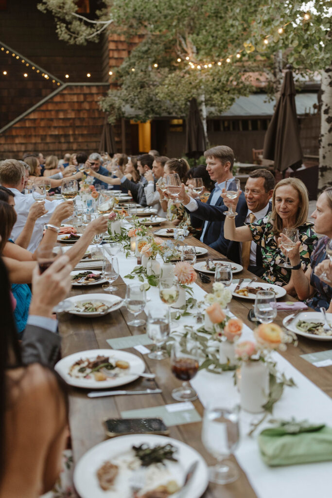Wedding guests holding up wine glasses to toast while sitting at reception table with colorful florals and white tableware at the PlumpJack Inn