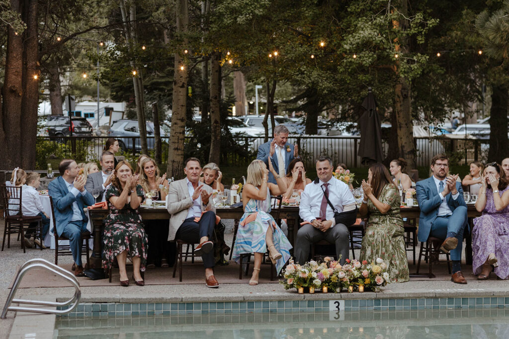 Wedding guests clapping while sitting at reception tables with colorful florals and string lights and trees in background