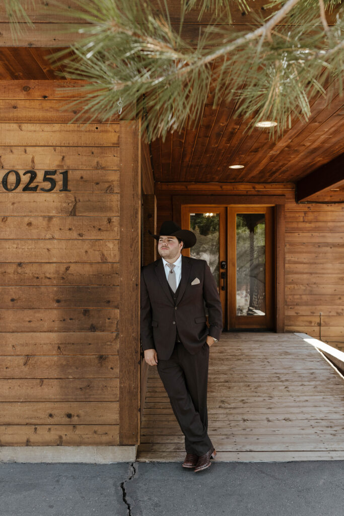 Wedding groom leaning on side of all wooden building with hand in pocket and looking of to the side at Austin Ridge Truckee