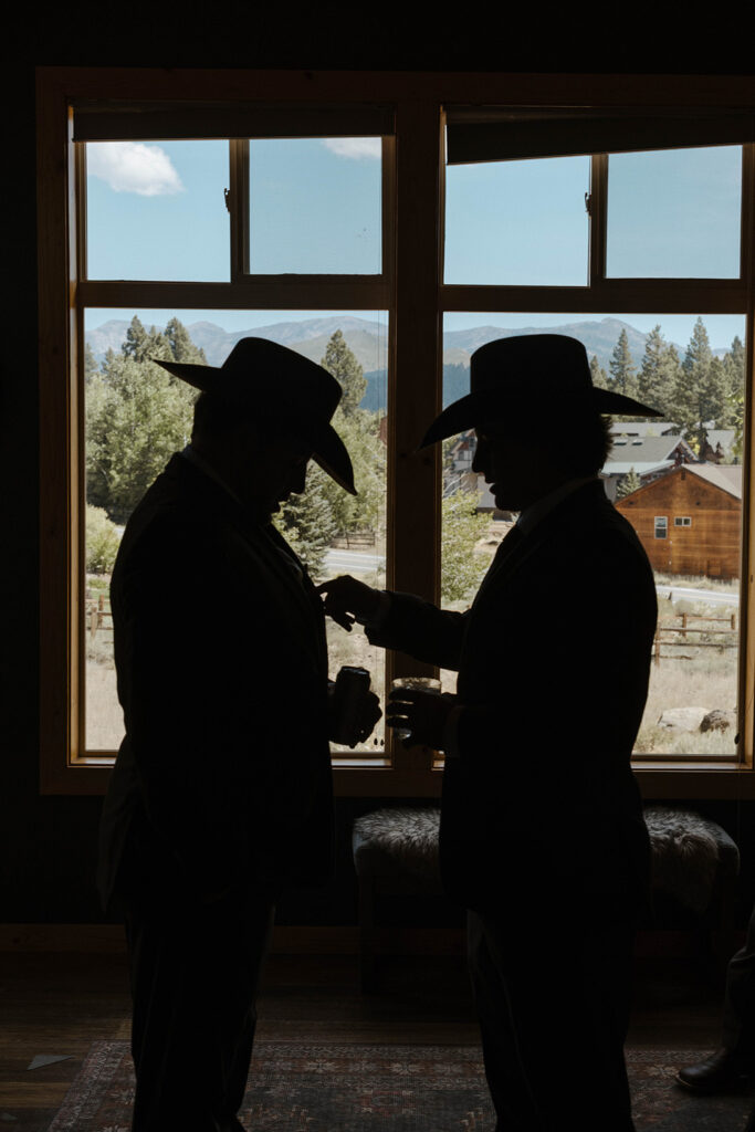 Silhouettes of wedding groom and groomsmen holding drinks together in front of windows while groomsmen adjusts groom's jacket at Austin Ridge Truckee