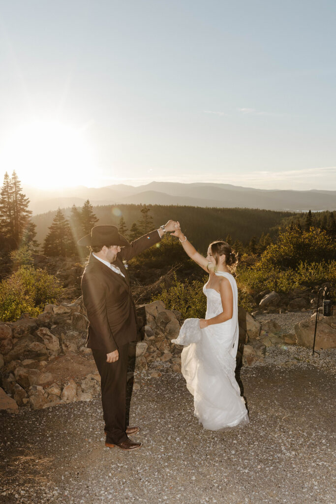 Wedding couple holding hands in air together while standing in front of rocks and shrubs with mountains and sunset in background at Austin Ridge Truckee