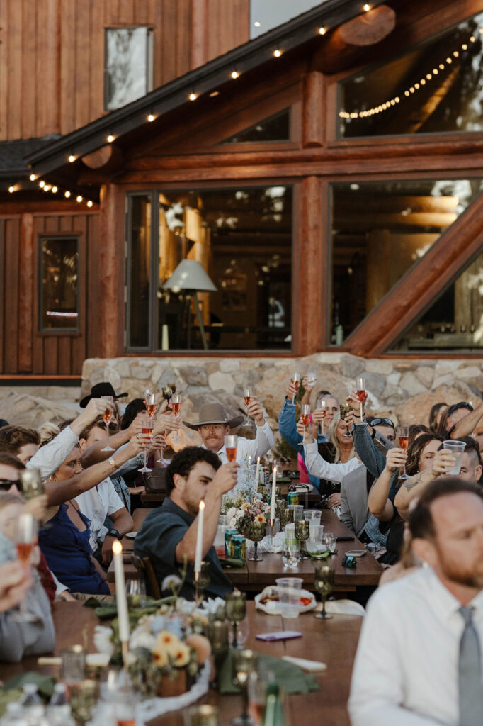 Wedding guests holding drink glasses up after speech during wedding reception with wooden building and rock wall in background at Austin Ridge Truckee