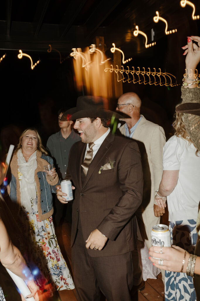 Groom laughing while holding beer during wedding reception with guests celebrating around him and lights blurring at Austin Ridge Truckee