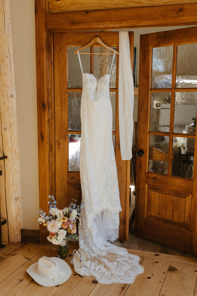 Wedding dress hanging on wooden door next to colorful flower bouquet and white cowboy hat at Austin Ridge Truckee