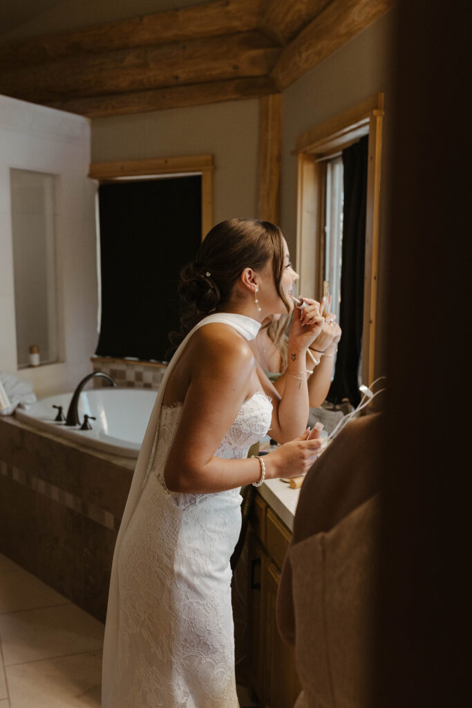 Bride looking in mirror while applying lipstick and wearing wedding dress inside at Austin Ridge Truckee