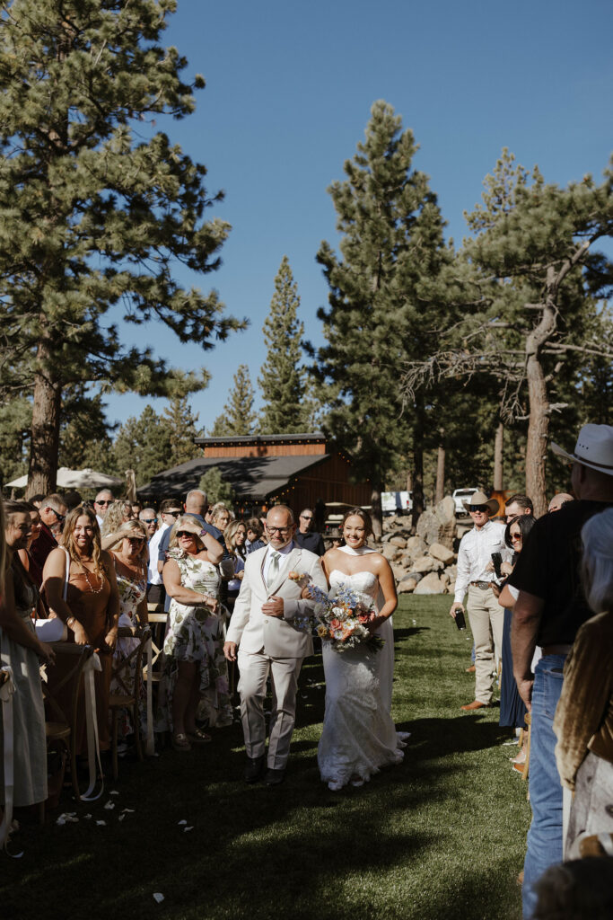 Bride holding dad's arm while walking down wedding aisle to ceremony at Austin Ridge Truckee with guests on either side and tall trees in background